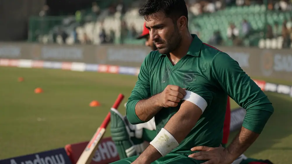 Close-up of a bearded cricketer in a green long-sleeve jersey adjusting a white band on his left upper arm during a grass training session with orange cones and a bat behind him